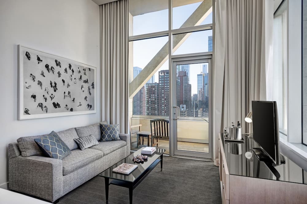 A sitting room with a grey sofa, glass coffee table, and tall windows opening to a balcony with city views.