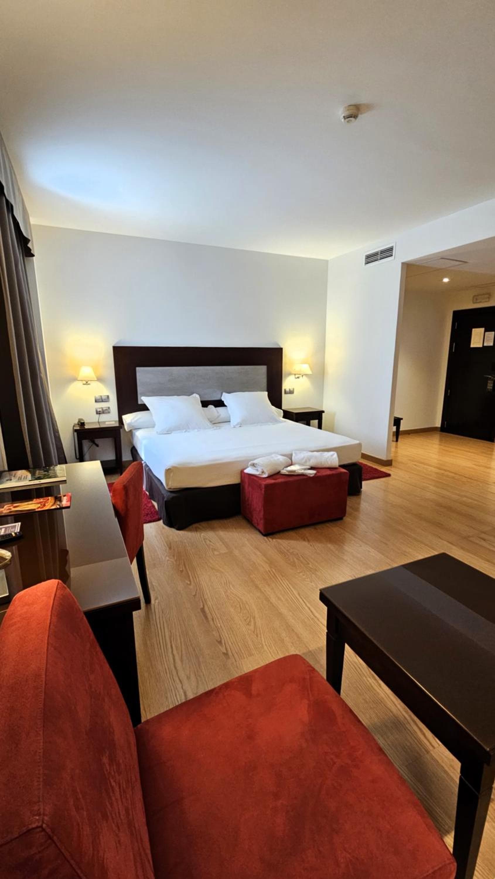 A hotel bedroom featuring wood flooring, red upholstered chairs, and a large bed with a dark headboard.