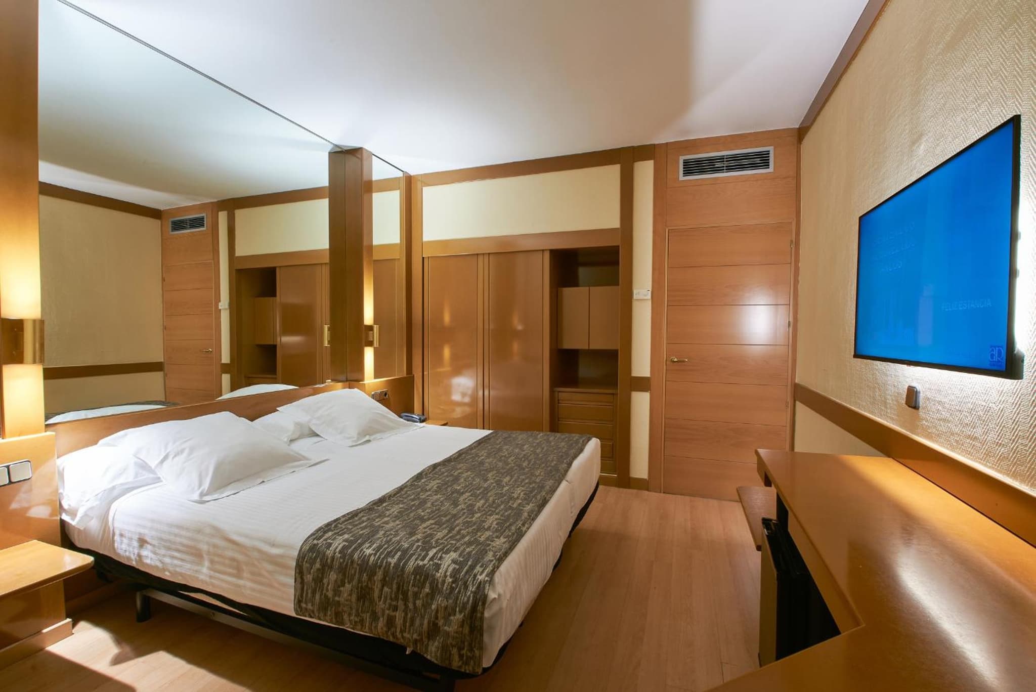 Hotel room featuring a made bed, wood wardrobe cabinetry, a mirrored accent wall, and a wall-mounted screen.