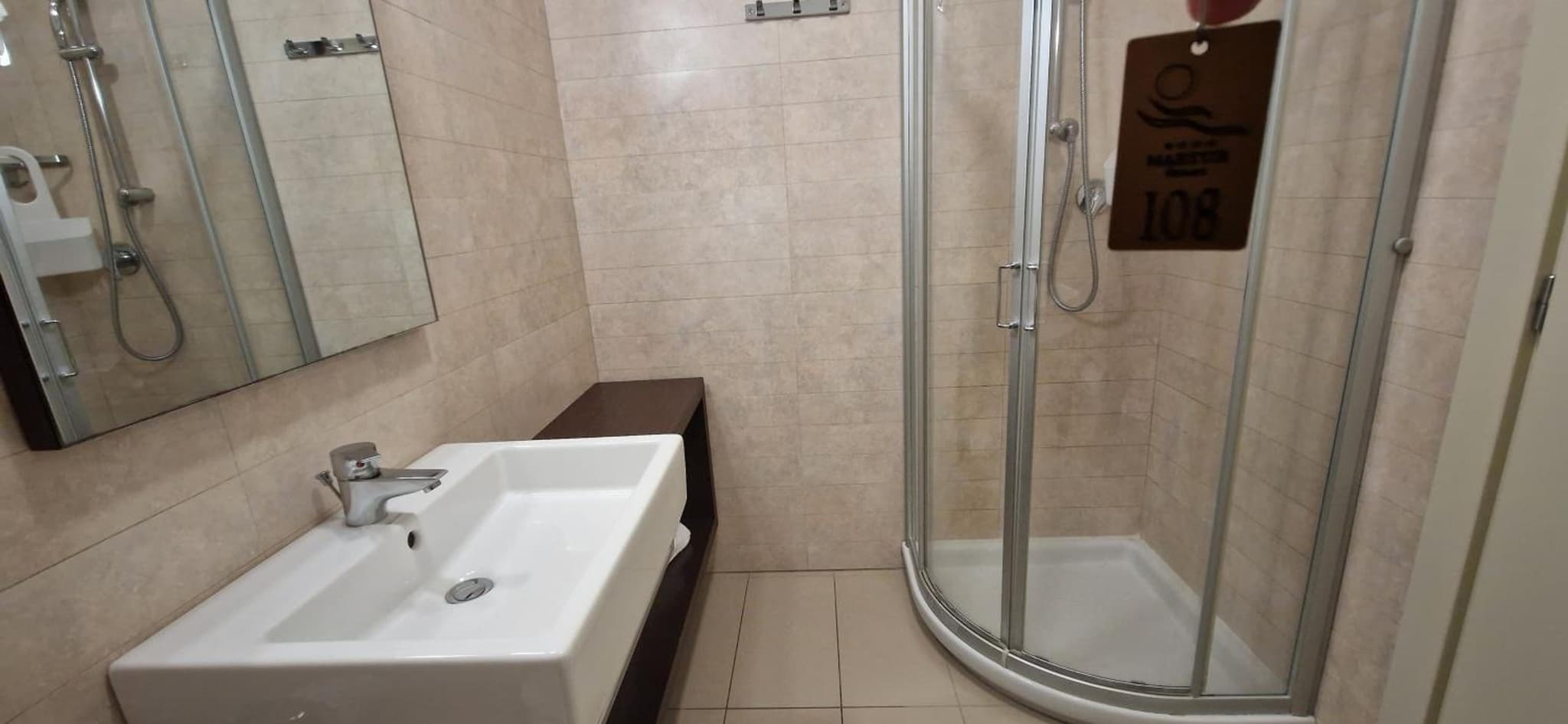 A modern bathroom with a white sink, glass corner shower stall, and light beige tiled walls.