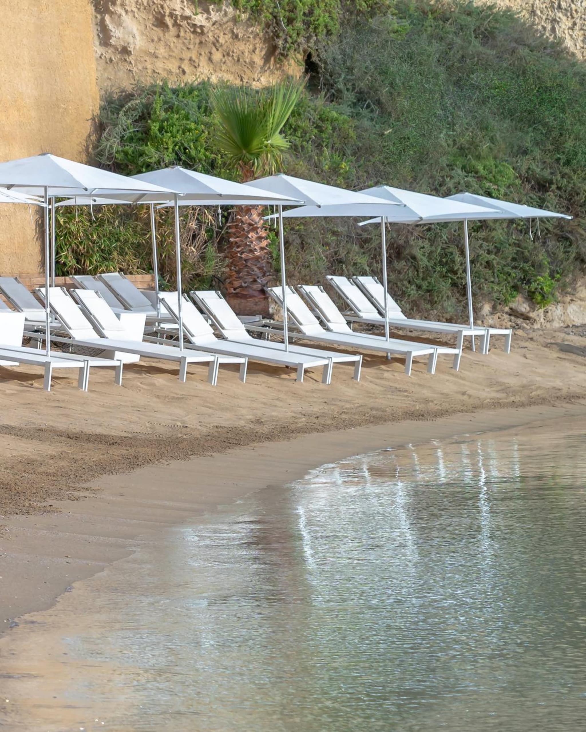 White sun loungers and square umbrellas set on the sand along the edge of calm coastal water.