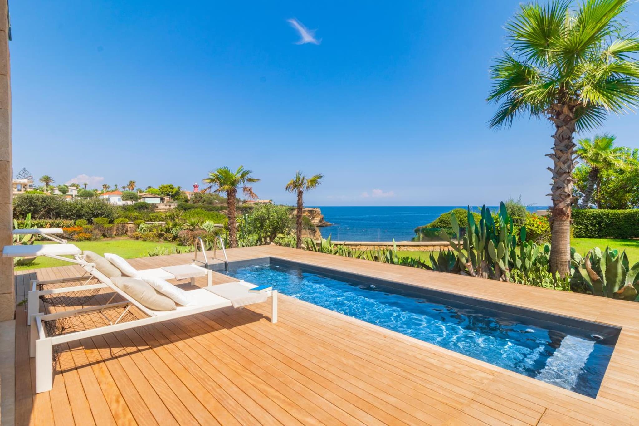 A plunge pool set into a wooden deck with sun loungers, surrounded by cacti, palm trees, and the blue sea beyond.