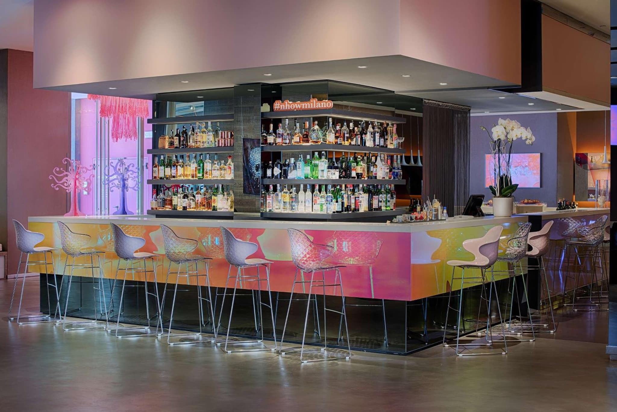 A contemporary bar with an iridescent front panel, high barstools, and illuminated shelves of bottles.