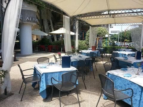 Outdoor dining patio under a sun canopy with blue tablecloths and dark metal chairs on gravel.
