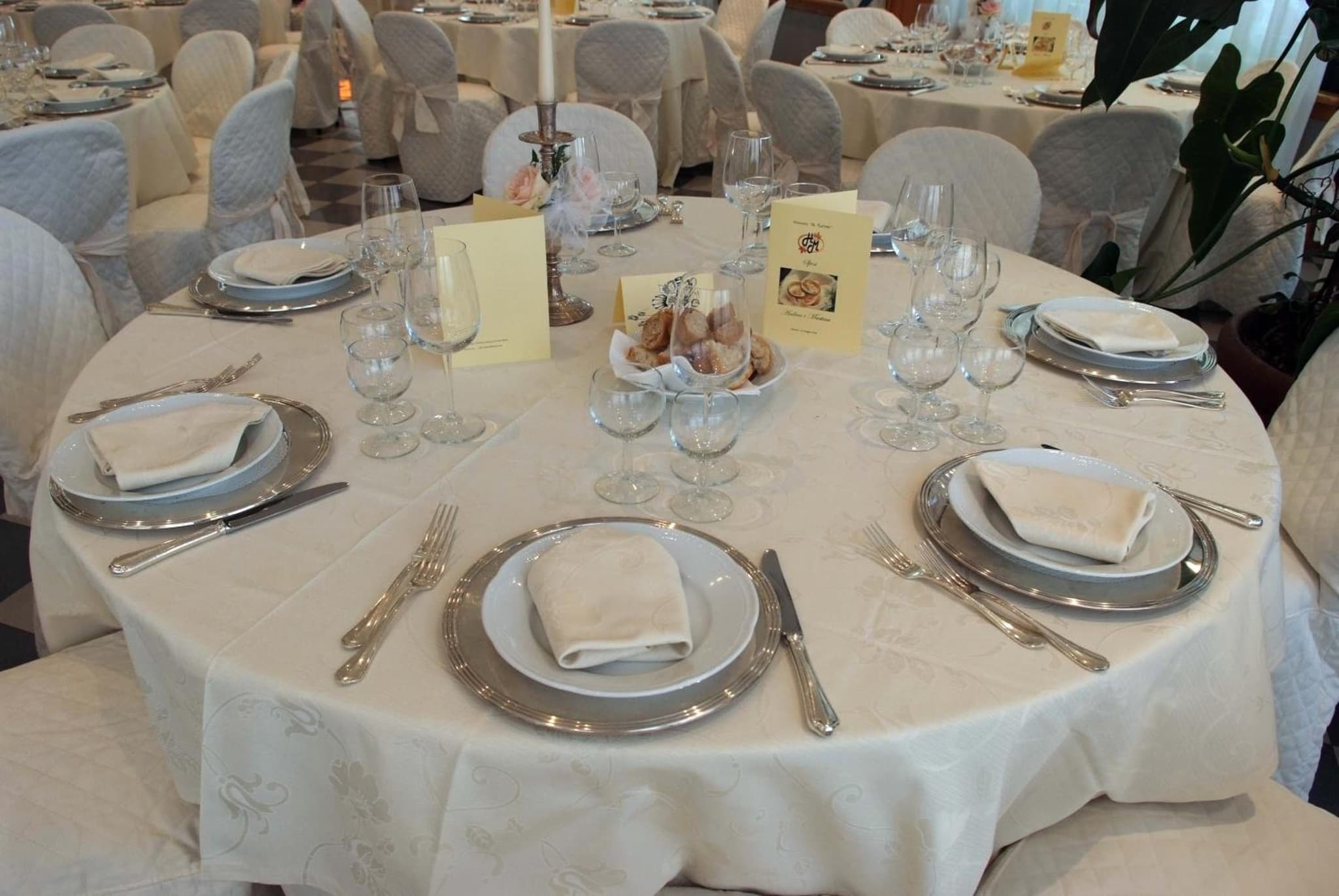 Formally set round dining table with white tablecloth, silver chargers, folded napkins, and stemware.