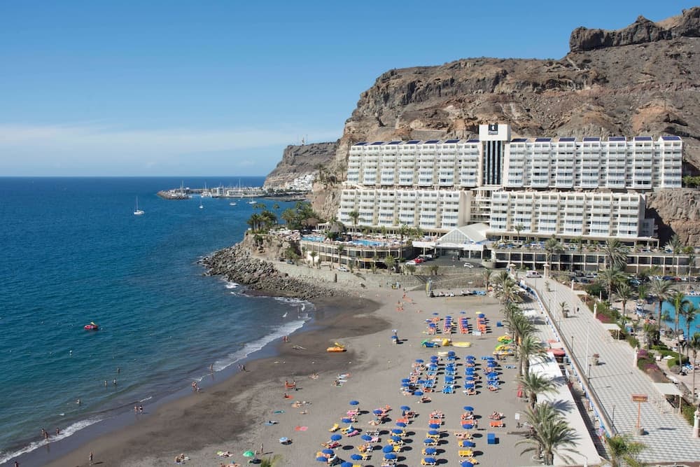 Elevated view of a dark sand beach with sun loungers fronting the resort against rugged cliffs and the sea.