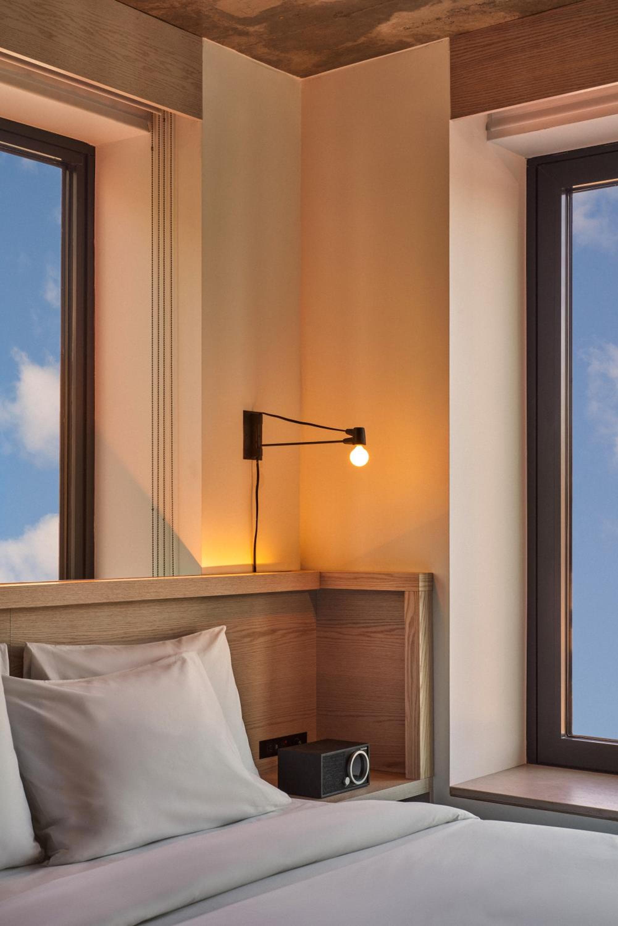 Hotel bedroom corner with a light wood headboard, bedside reading lamp, and windows framing a blue sky.