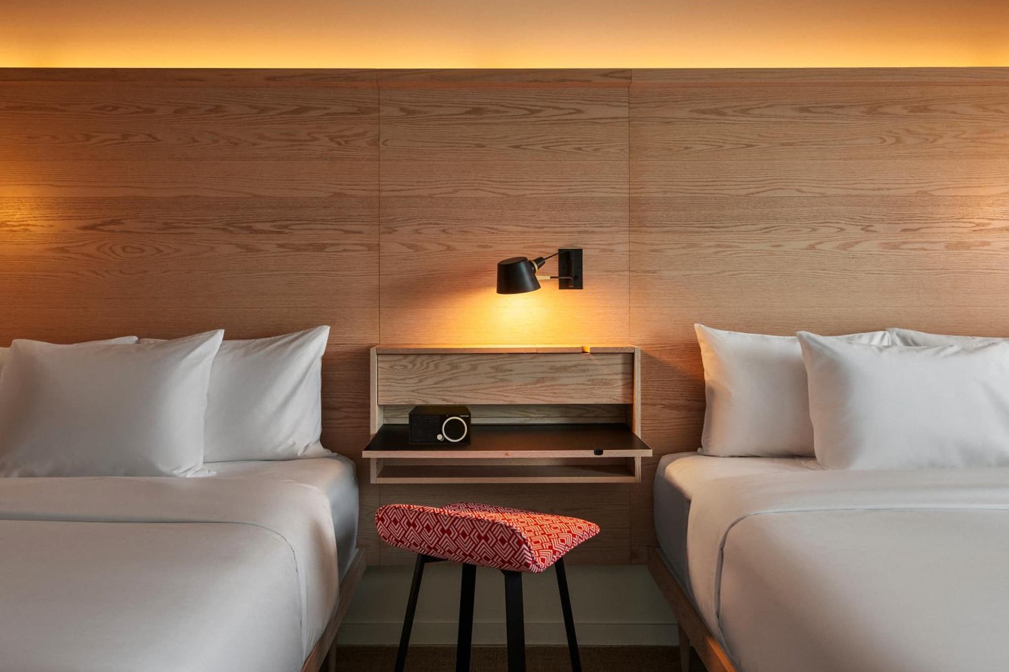 Hotel bedroom featuring beds with white linens flanking a wood panel wall with a sconce and floating shelf.