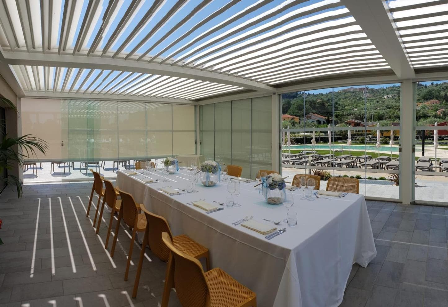 A long banquet table set with white linen under a louvered pergola enclosing a terrace with glass walls.