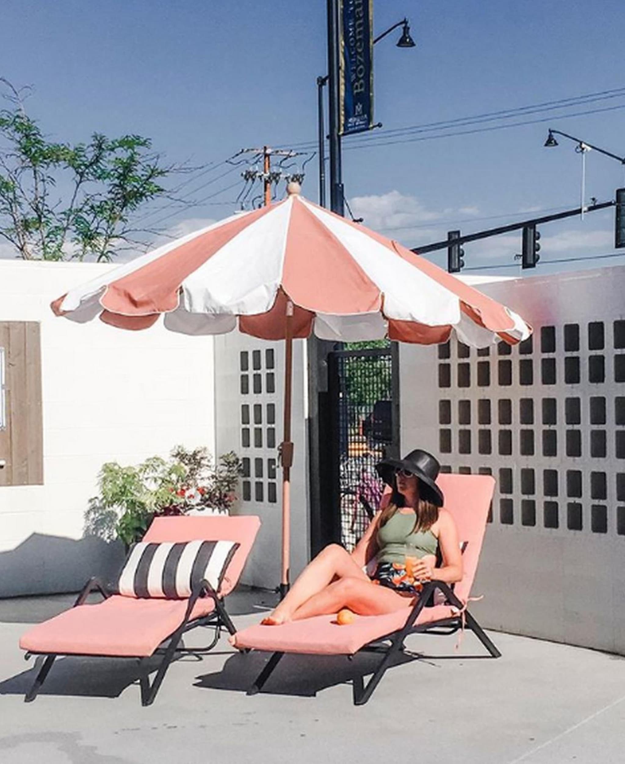 A woman relaxes on a pink sun lounger beneath a striped umbrella beside a breeze block wall.