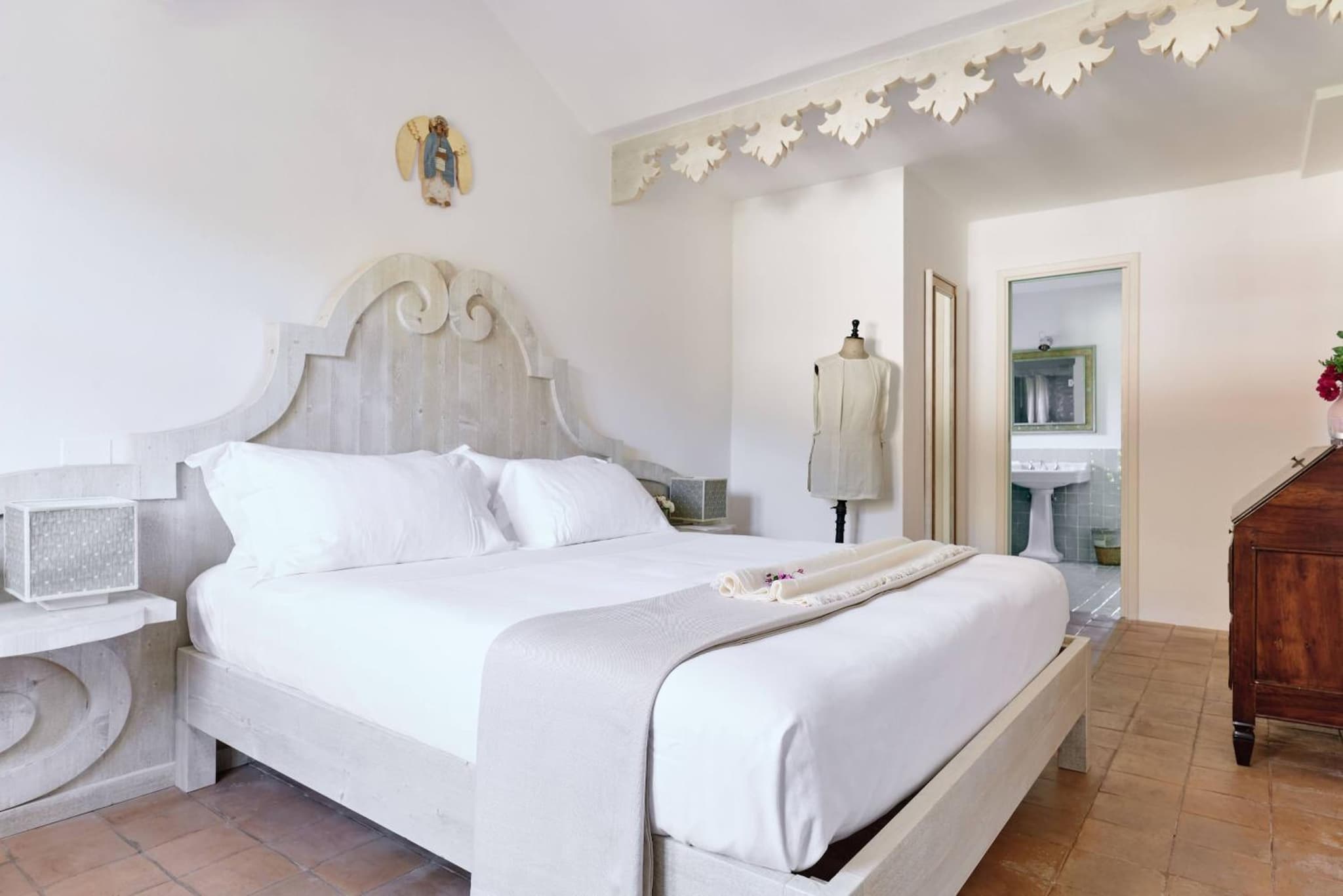 A bedroom with a carved wooden headboard, white linens, terracotta floor tiles, and a doorway to a bathroom.