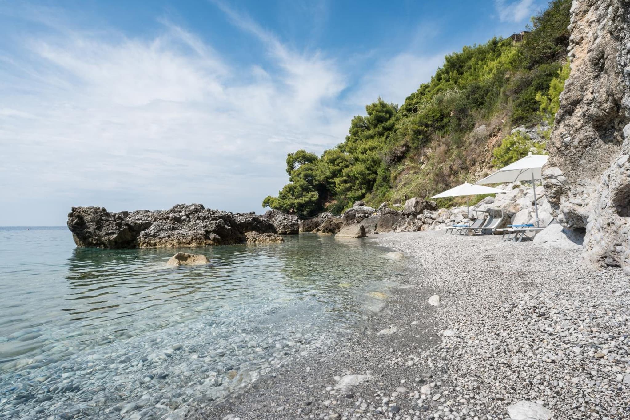 A pebble beach with clear water, sun loungers beneath umbrellas, and rocky cliffs covered in green trees.