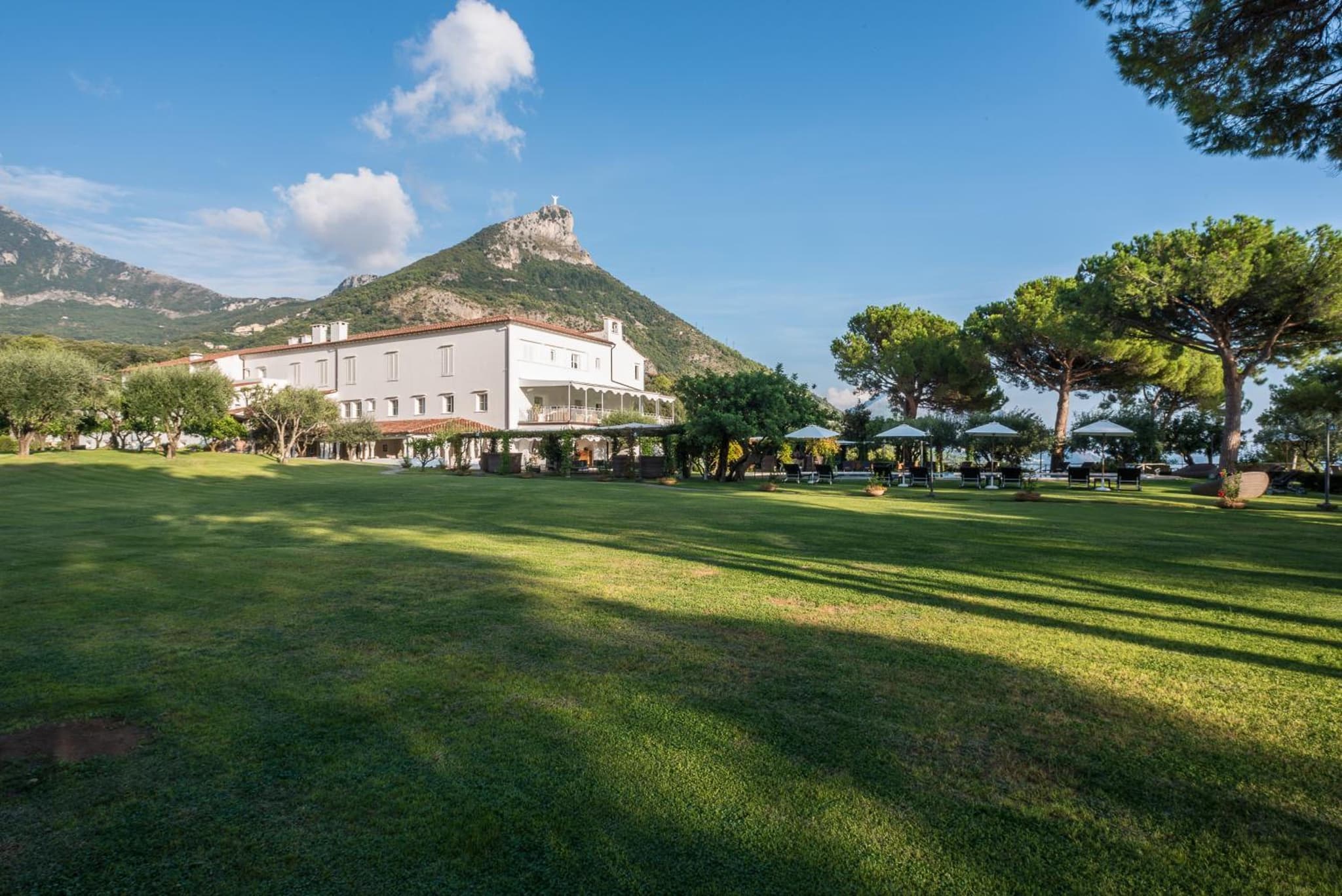 A wide green lawn facing the hotel facade with a rocky mountain peak rising behind under blue skies.