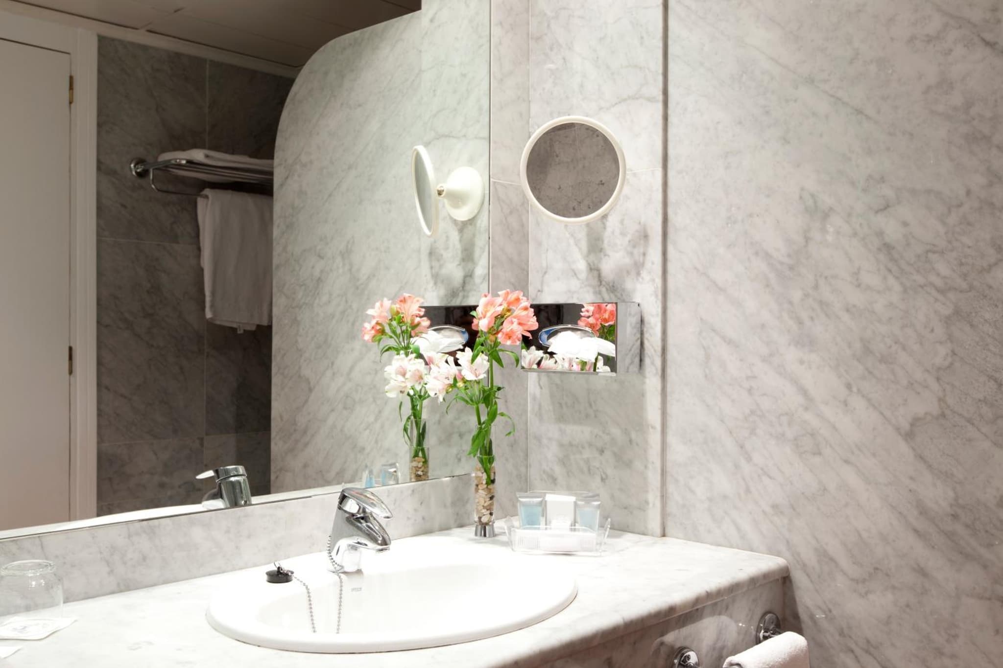 A marble bathroom vanity with an undermount sink, vase of flowers, and wall-mounted magnifying mirror.