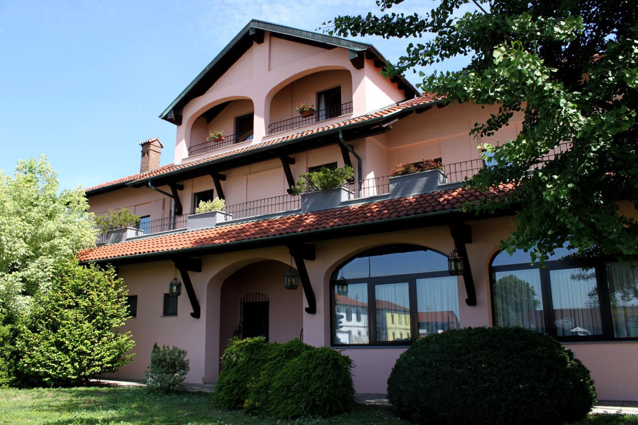 The pink facade features arched upper balconies, terracotta roof eaves, large windows, and manicured green shrubs.