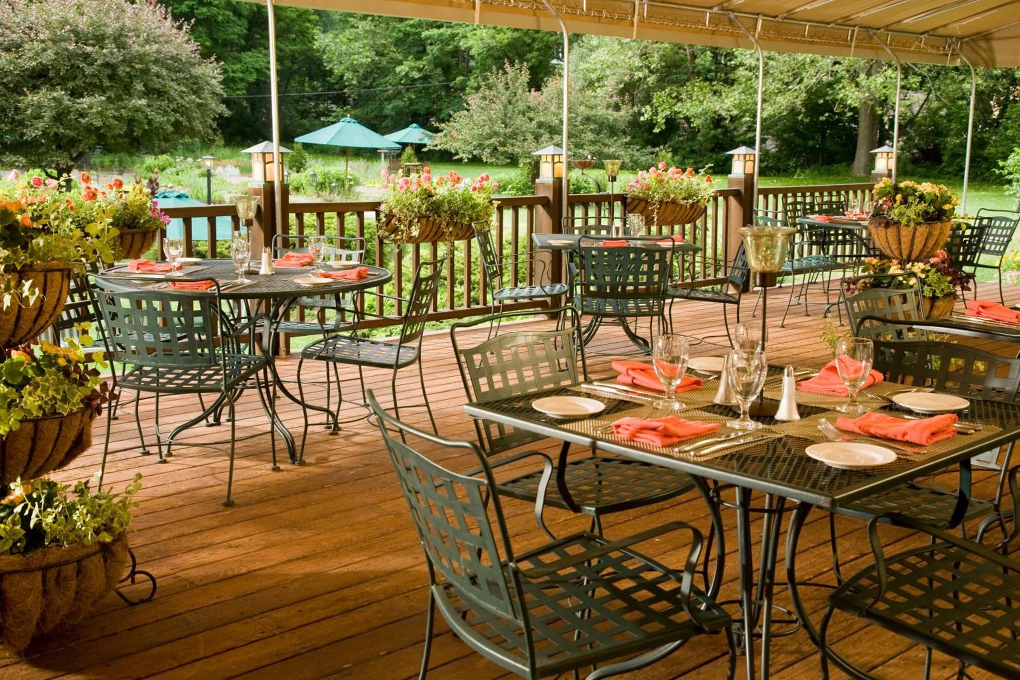 Covered wooden deck set with metal dining tables, glassware, folded napkins, and hanging flower baskets against trees.