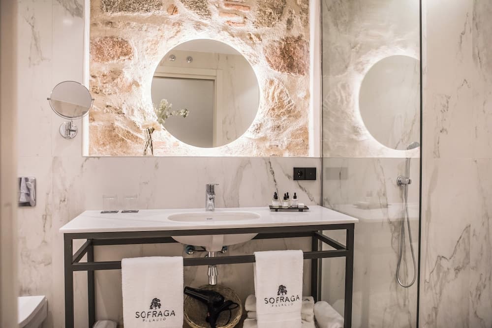 Modern bathroom featuring a marble vanity, exposed stone wall, and a circular backlit mirror next to a shower.