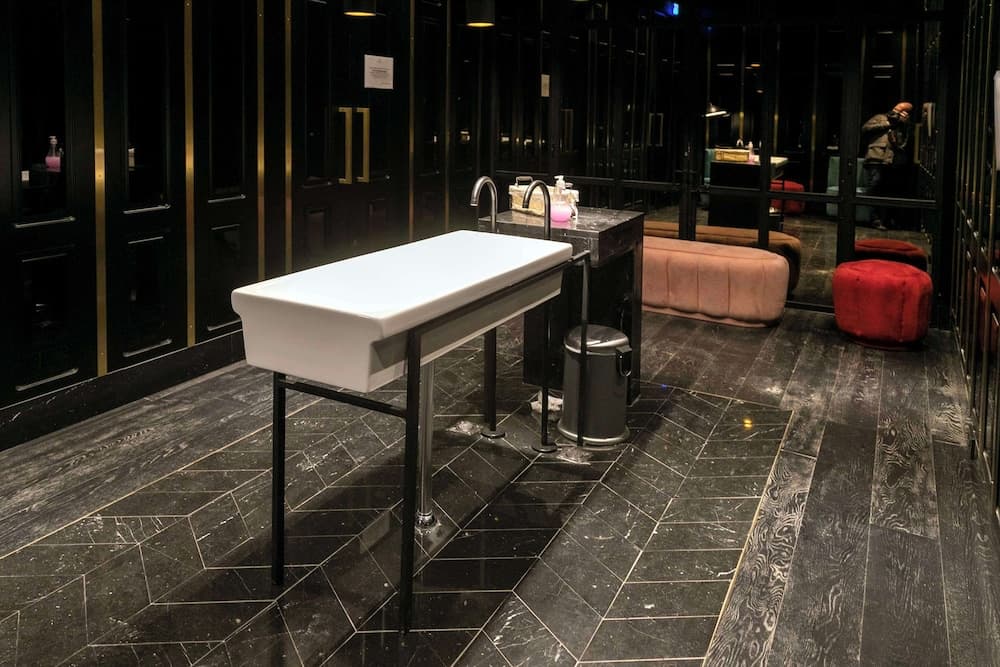 Restroom with dark wood-grain chevron tile floors, black paneled walls, and a white trough sink on metal legs.