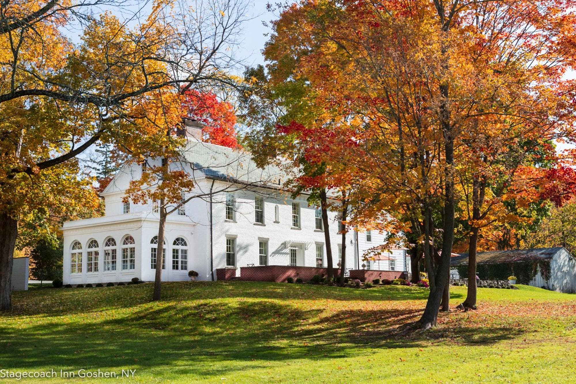 The white building seen across a grassy lawn shaded by trees with red, yellow, and orange autumn leaves.