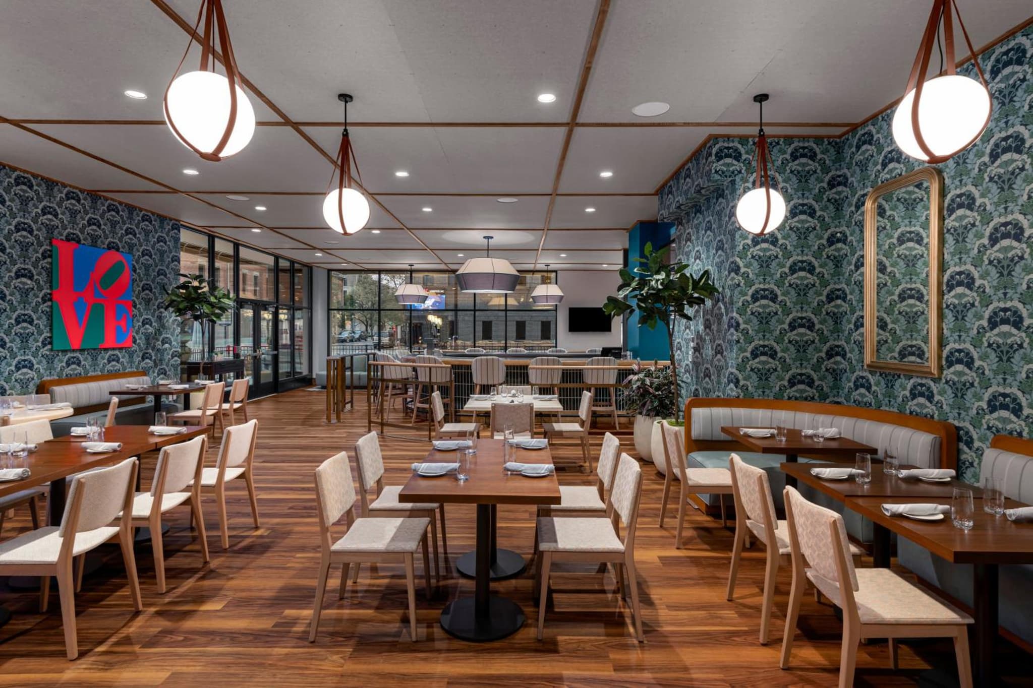 Restaurant dining room with botanical wallpaper, wooden tables and chairs, booth seating, and globe pendant lamps.