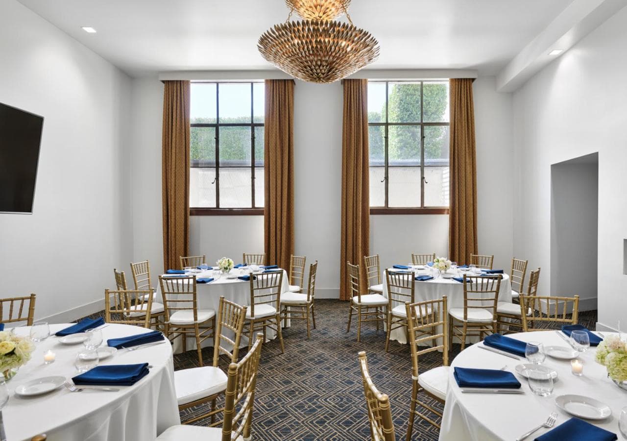 Event dining room with round tables covered in white linens, gold chairs, tall windows, and a metallic chandelier.