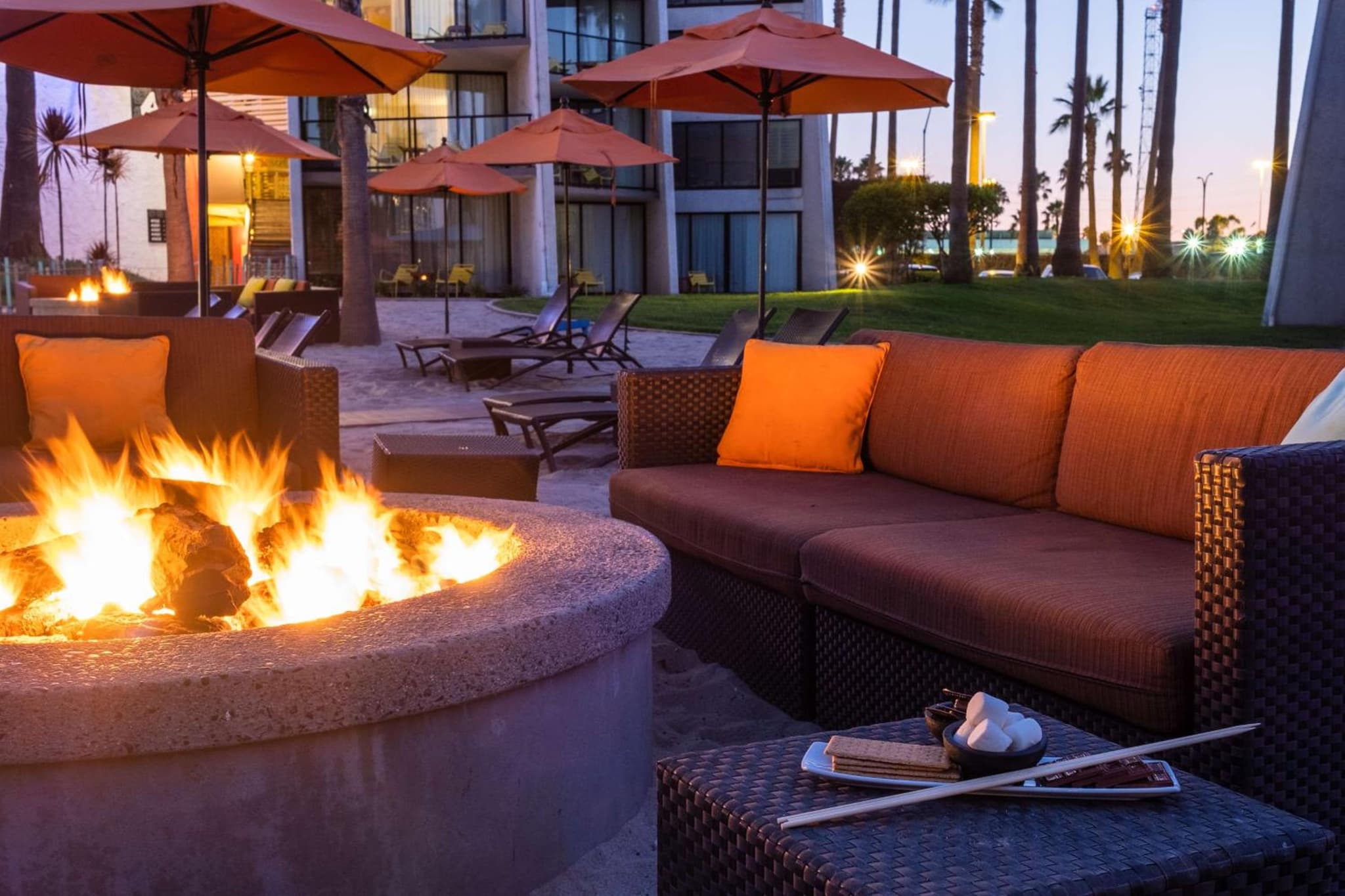 A flaming stone fire pit surrounded by wicker patio seating with orange cushions and umbrellas at twilight.