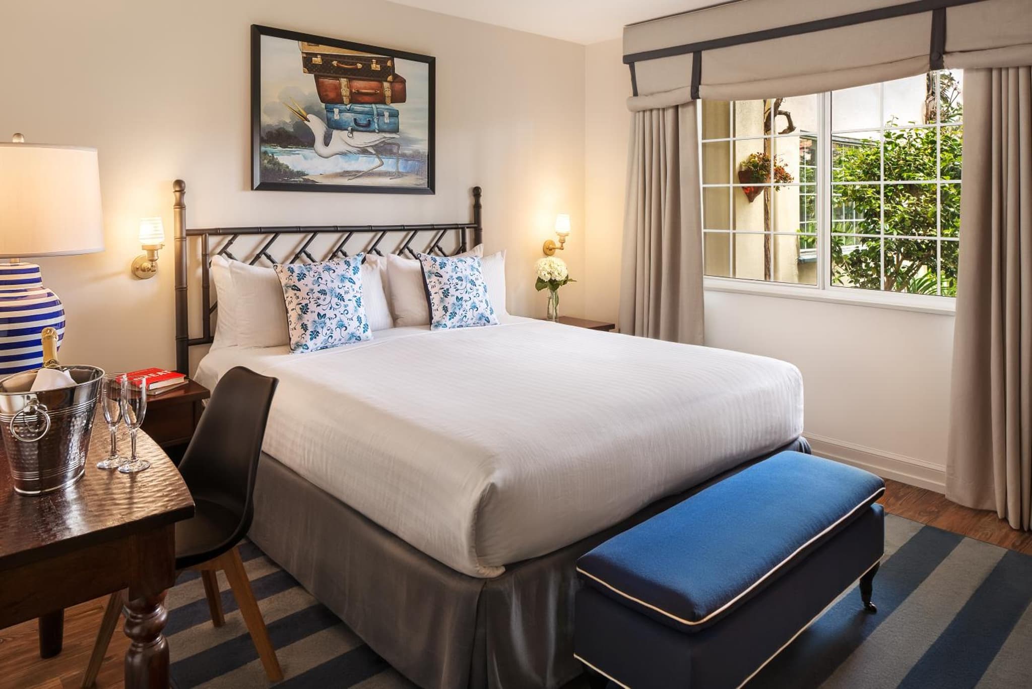 A bedroom featuring an iron headboard, white bedding, an upholstered blue bench, and a window to courtyard greenery.