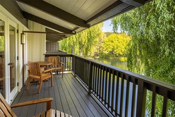 A covered wooden balcony with outdoor seating overlooks a pond bordered by weeping willow trees.