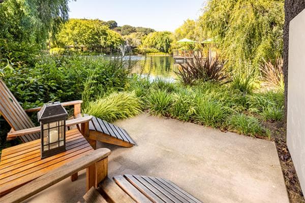 A concrete patio with wooden lounge chairs overlooks landscaped garden beds and a pond.