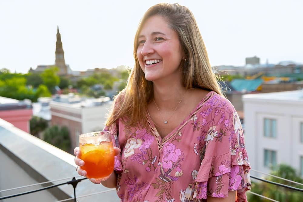 A woman smiles holding a cocktail on a rooftop terrace overlooking the city skyline under daylight.