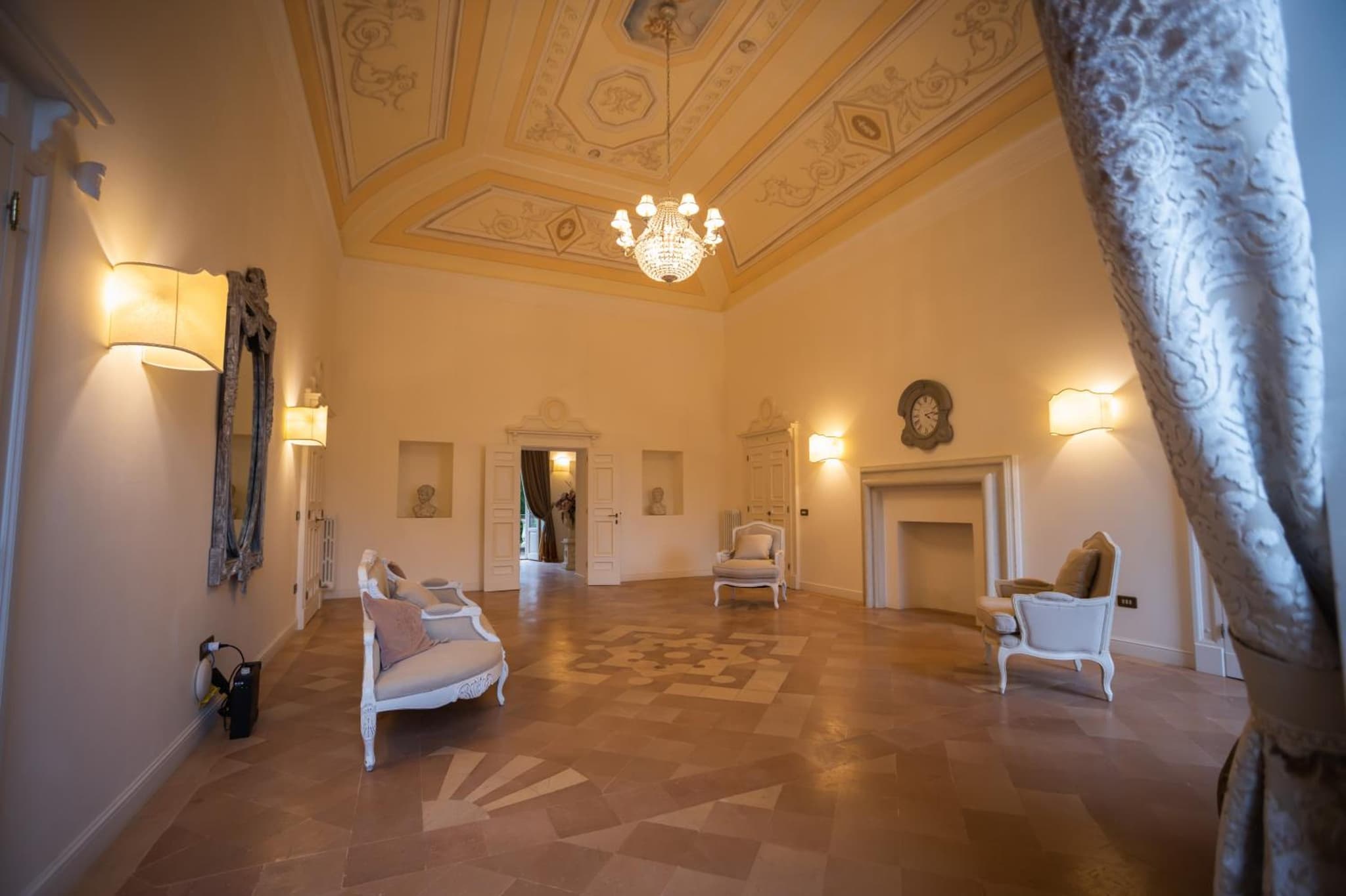 A formal sitting room featuring ornate ceiling frescoes, a chandelier, a stone fireplace, and antique seating.