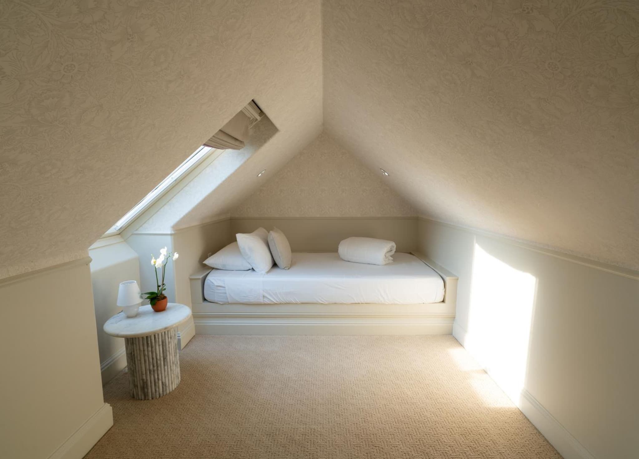 A bed is built into an alcove beneath the pitched ceiling and skylight of a wallpapered attic room.