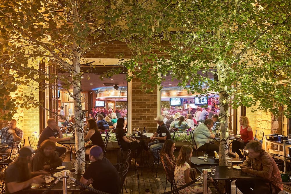 Diners sit at patio tables beneath string-lit birch trees outside a warm, window-lined restaurant.