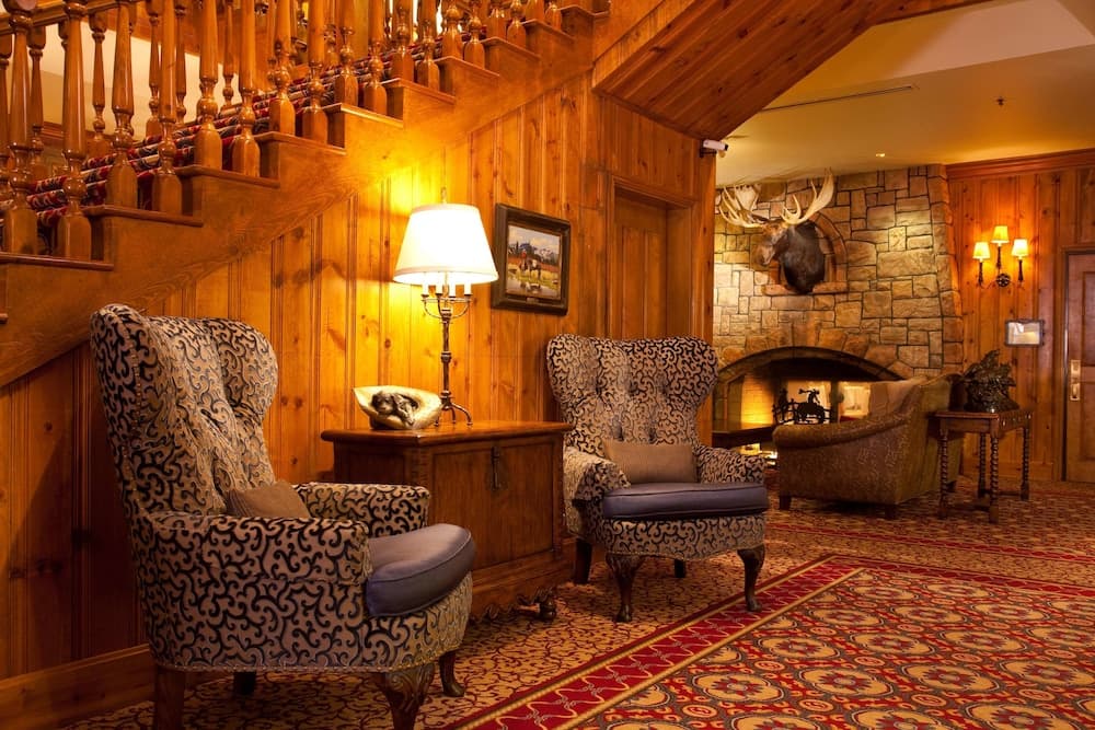 A wood-paneled lounge features patterned armchairs, an ornate rug, and a stone fireplace with a mounted moose head.