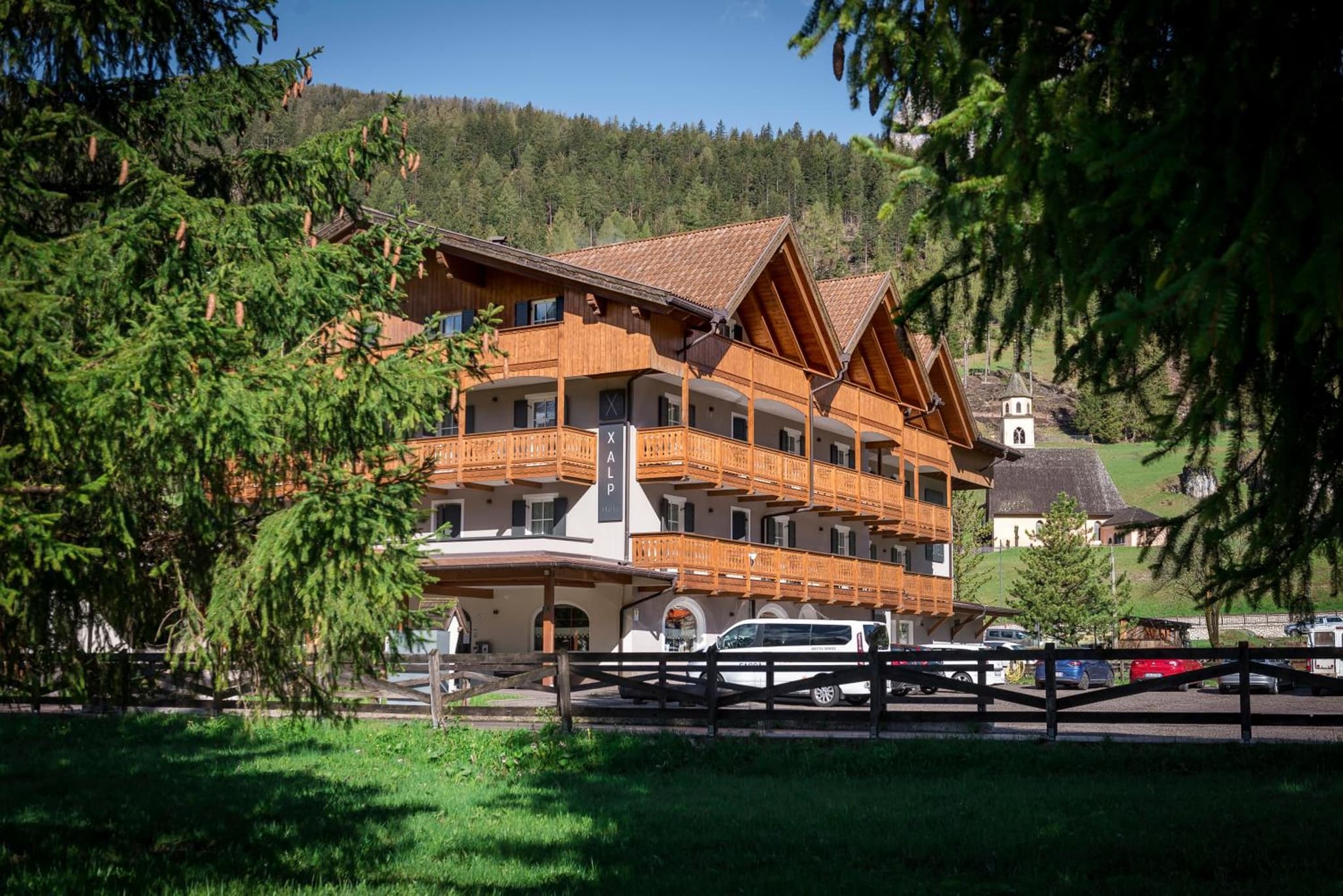 Daytime view of the timber alpine exterior framed by pine branches, with a village church and hills behind.