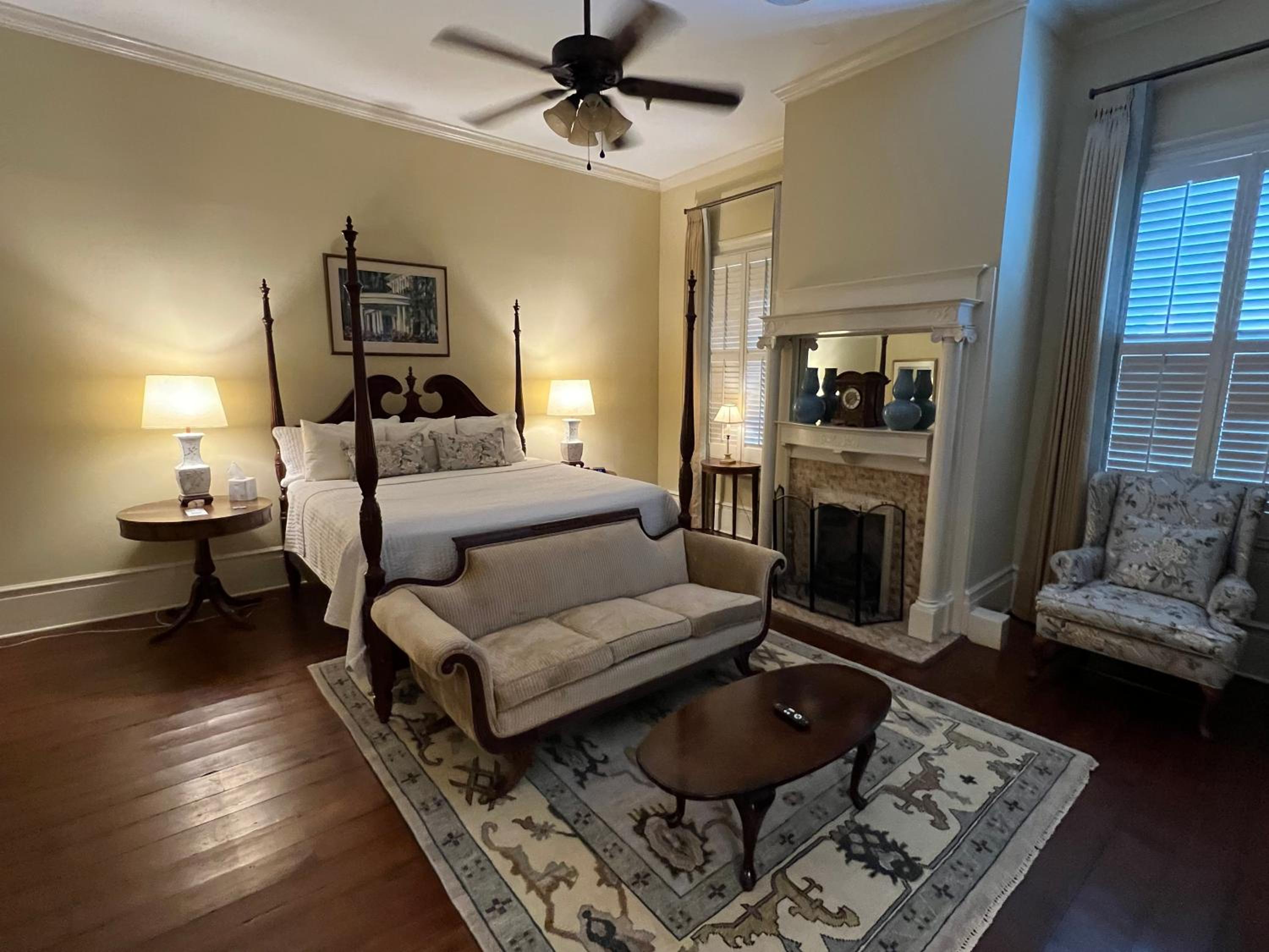 Bedroom with a tall wooden bed, an upholstered settee, polished wood floors, and a fireplace mantel.