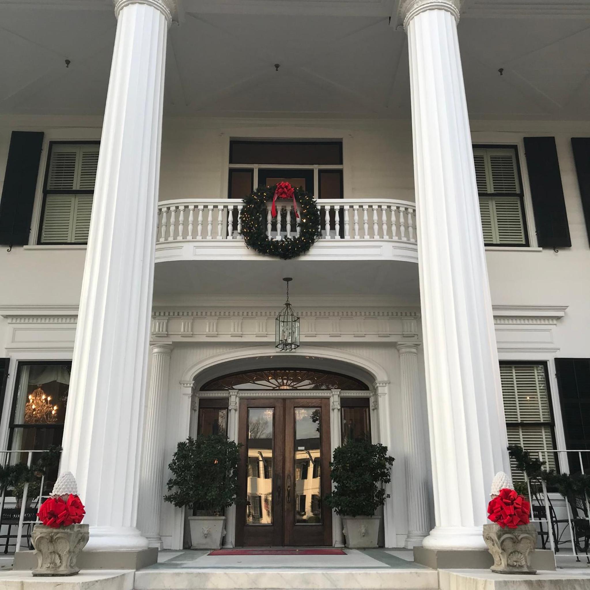Mansion portico featuring fluted columns, wooden entry doors, and a holiday wreath on the upper balcony.