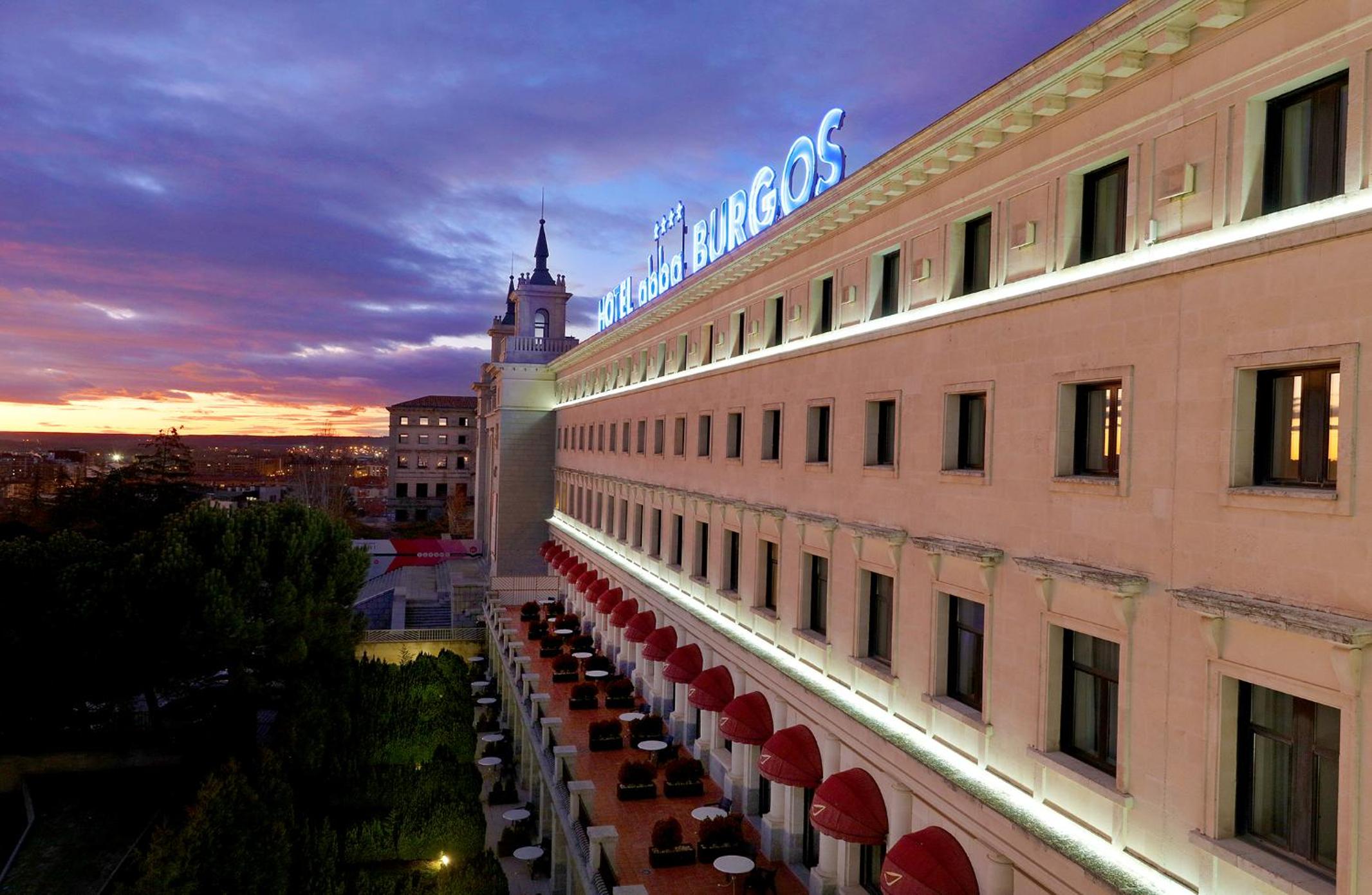 The stone exterior facade of Hotel Abba Burgos illuminated with glowing blue signage under a sunset sky.