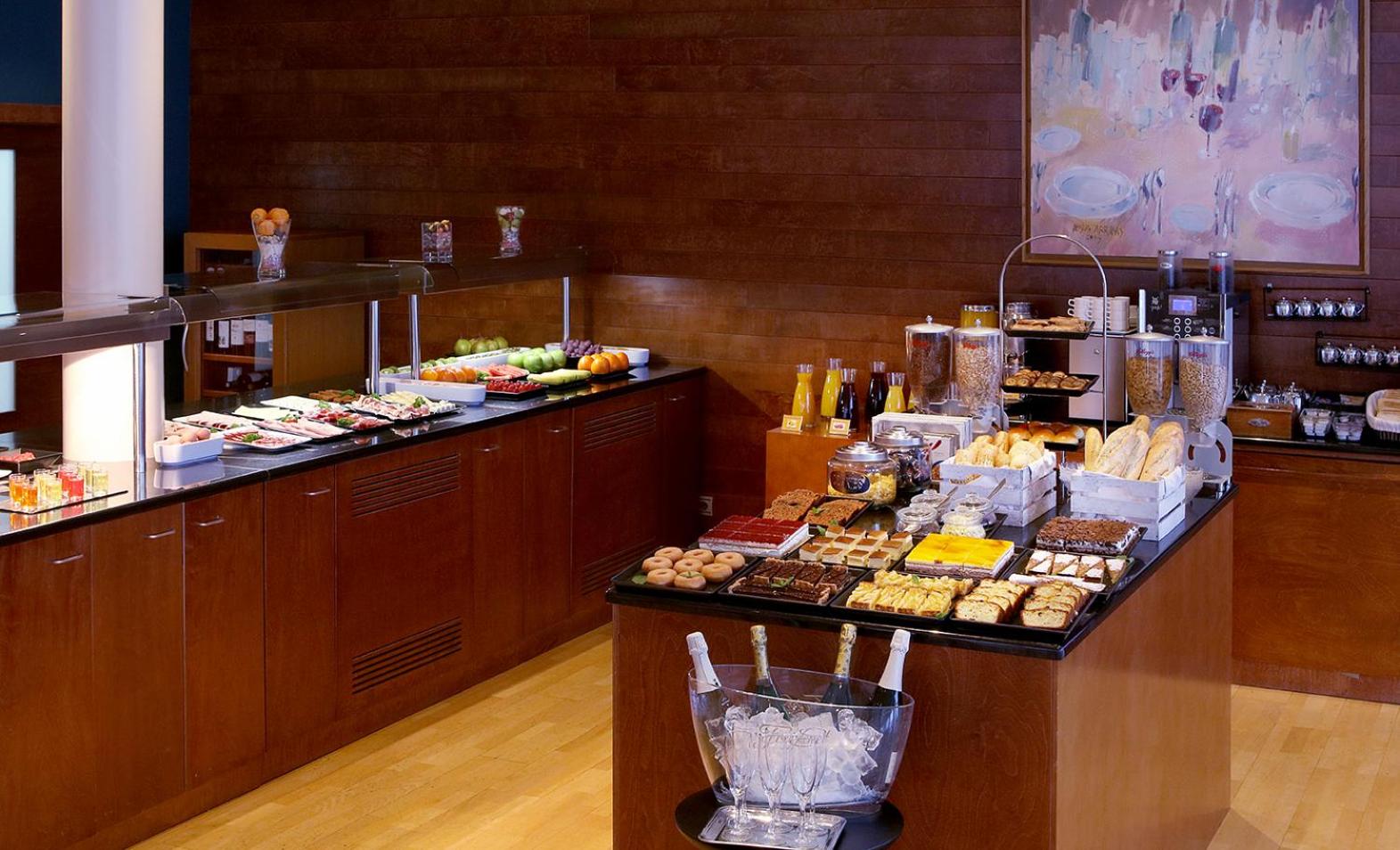 A wooden buffet counter displaying bread, pastries, fruit, and cereal beside chilled bottles on a stand.