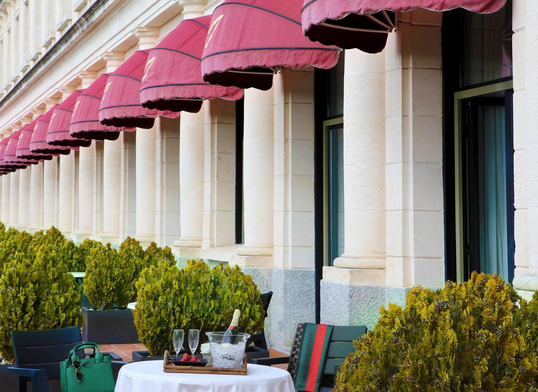 A colonnaded stone terrace beneath red awnings, with trimmed shrubs and a small table set with champagne.