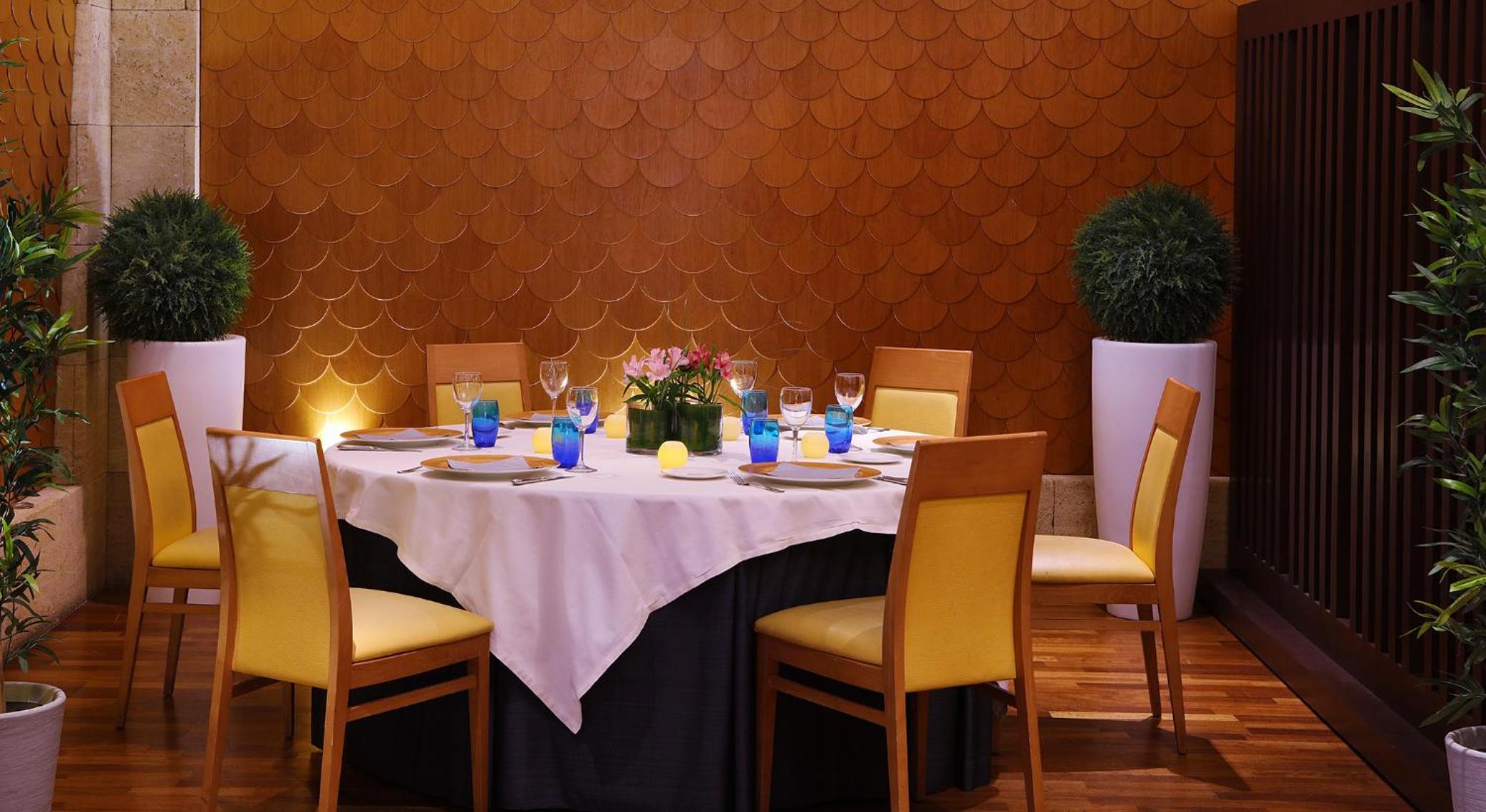 A round dining table set with white linen and blue glassware against a scalloped wood wall.