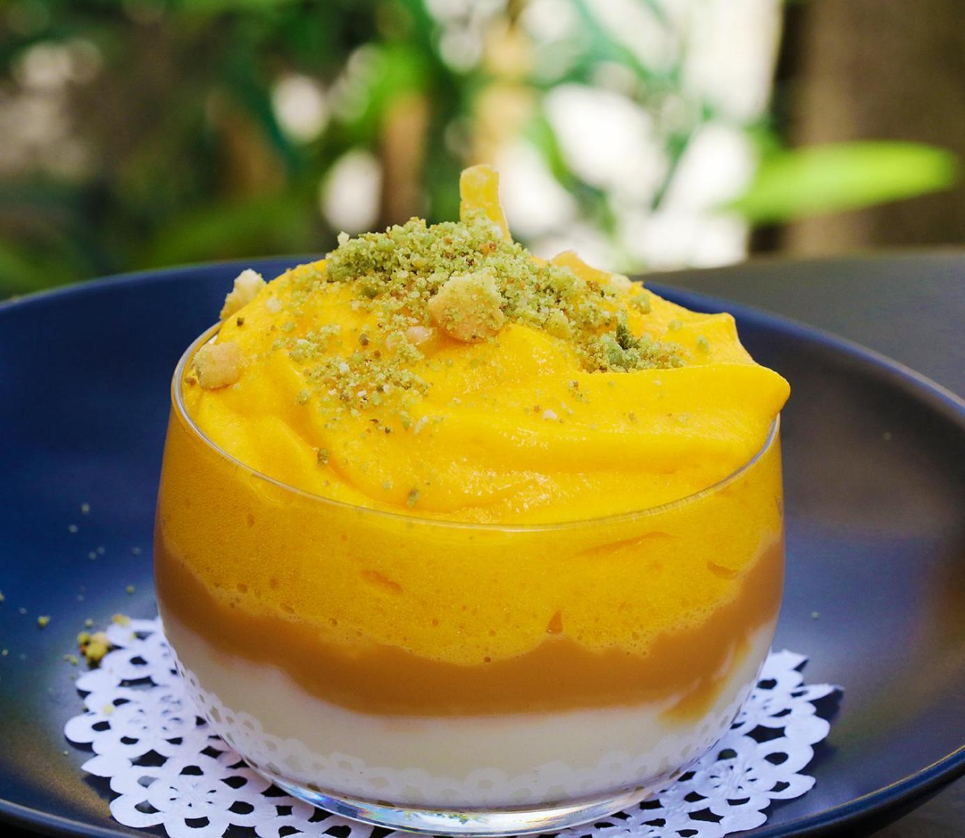 A layered dessert cup with white cream, caramel, and yellow mousse dusted with crushed nuts.