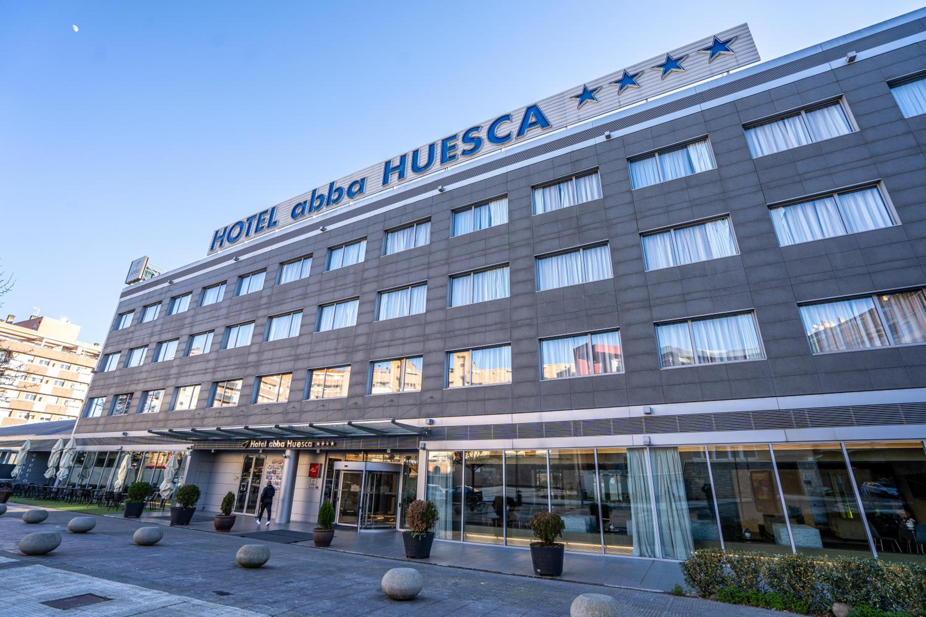 Facade of Hotel abba Huesca with grey tiled walls, large windows, and a glass entrance under clear daylight.