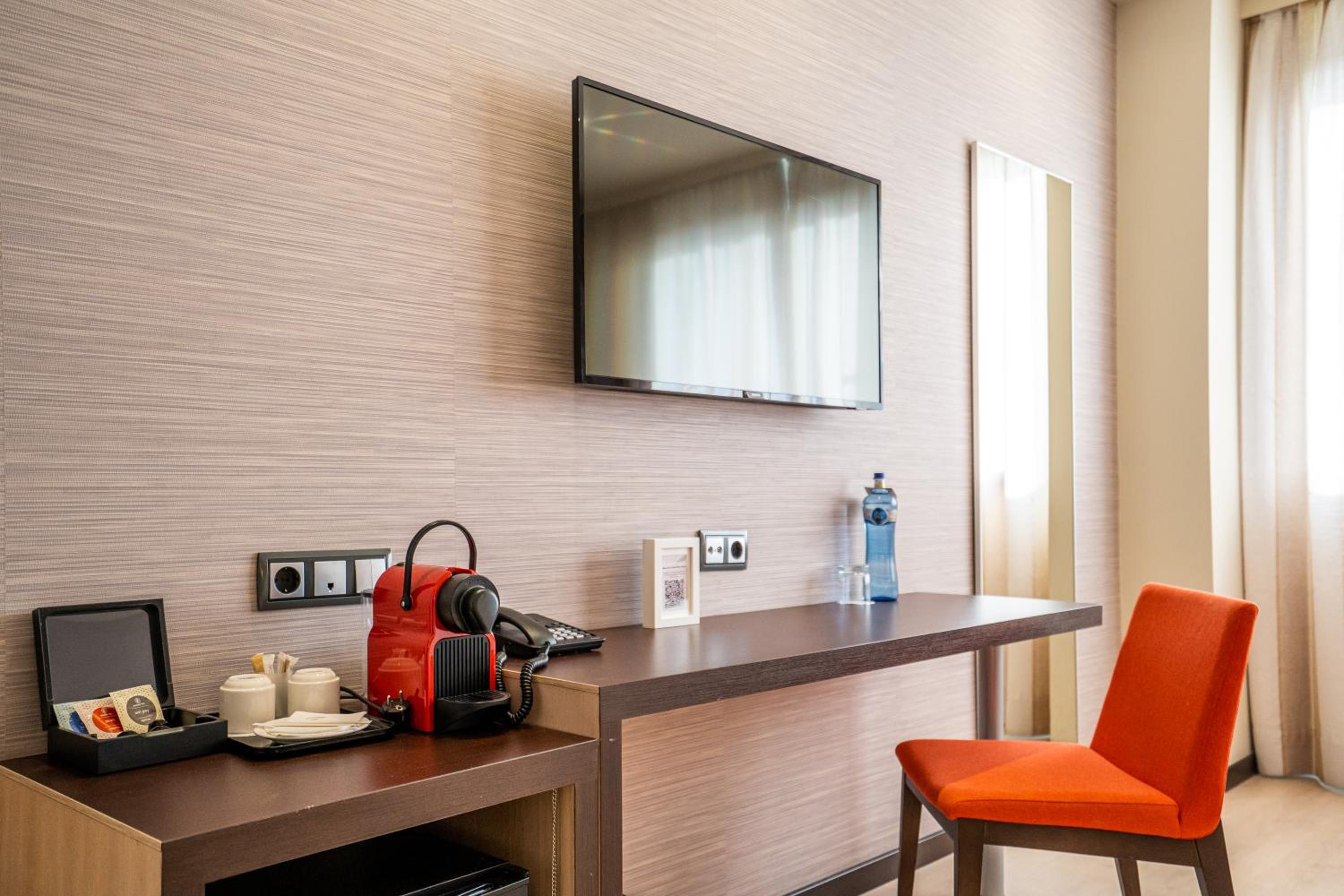 Wooden desk in a guest bedroom with an espresso maker, wall-mounted television, and an orange upholstered chair.