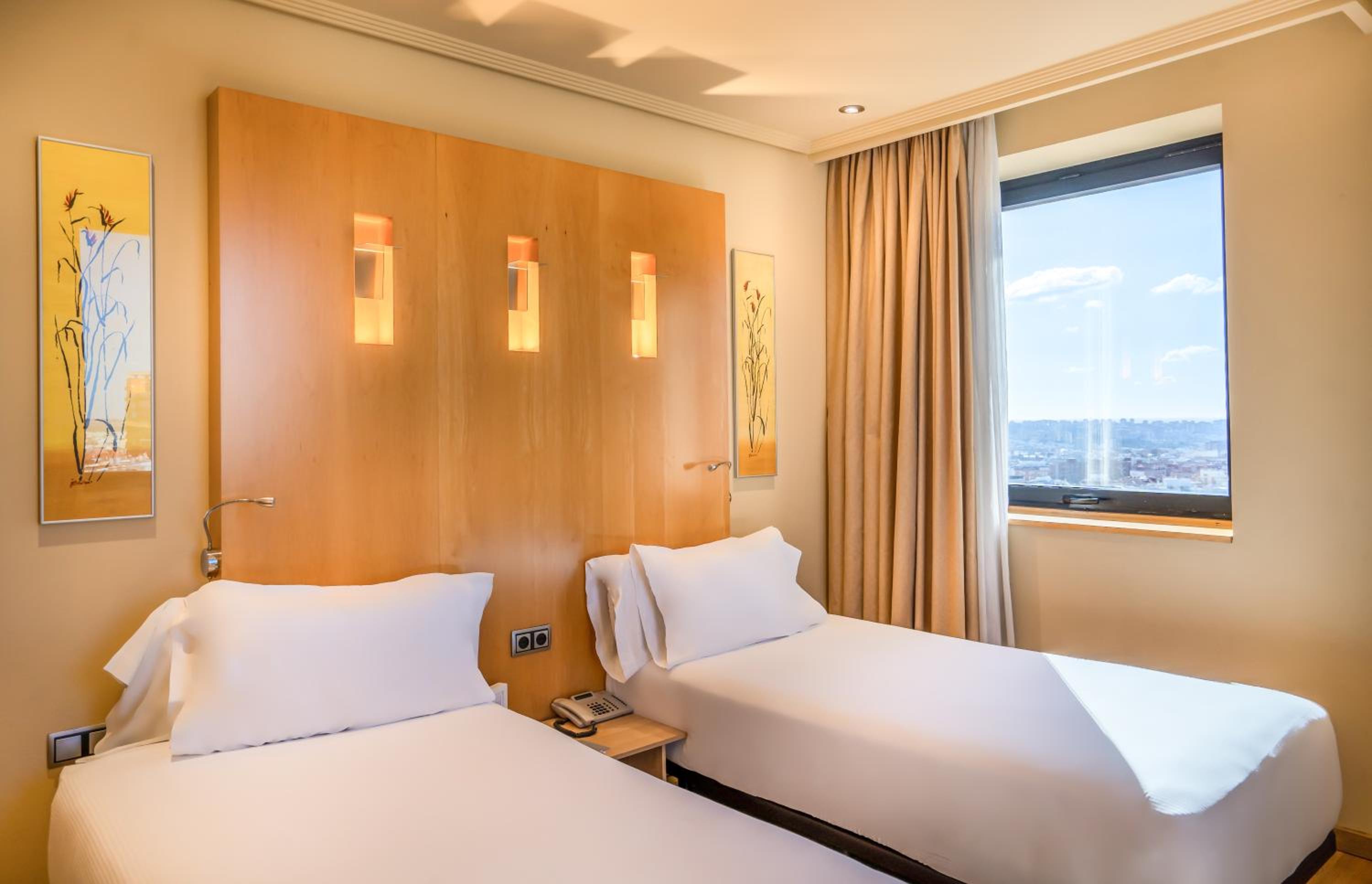 A hotel guest room with white-sheeted beds, a wooden headboard with recessed lighting, and a city-view window.