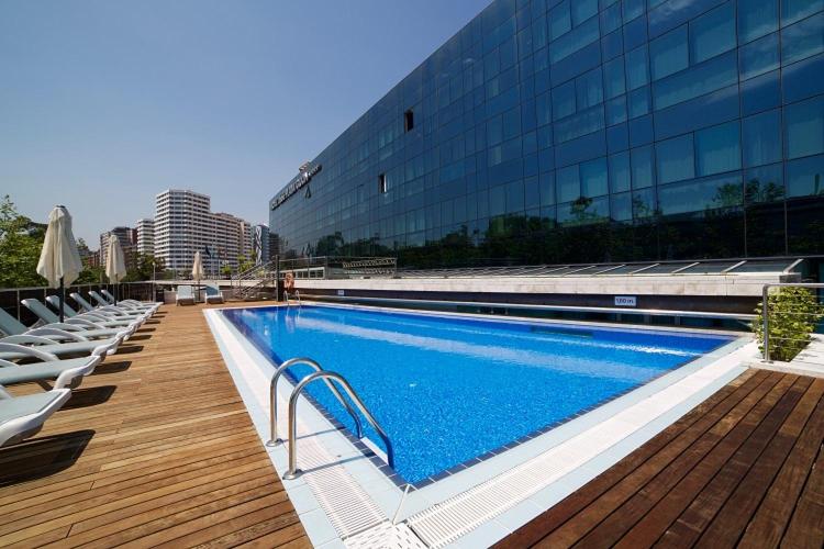 An outdoor swimming pool on a wooden sun deck beside the glass facade of Abba Playa Gijón.