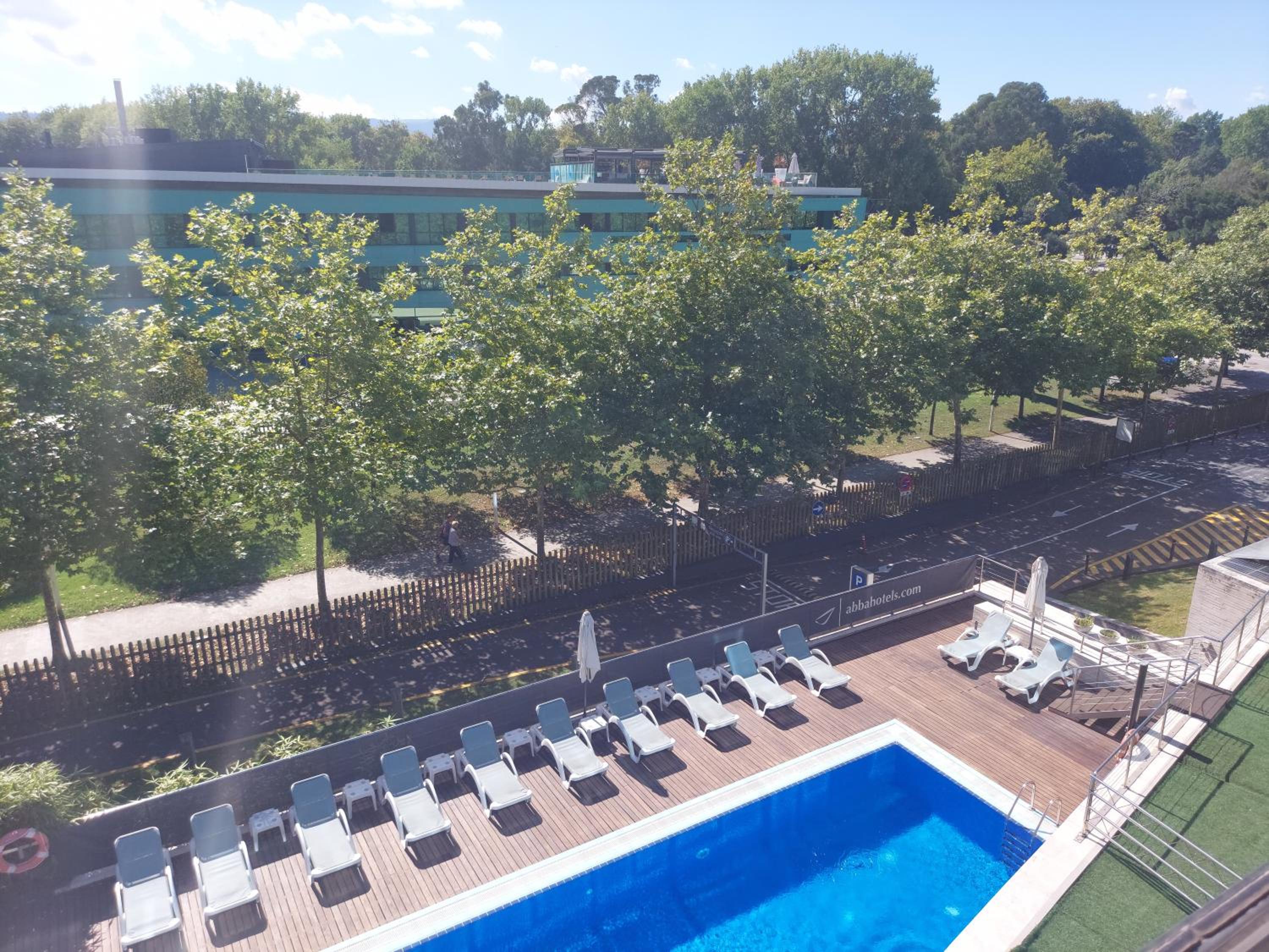 An elevated view of an outdoor swimming pool bordered by a wooden sundeck, lounge chairs, and green trees.