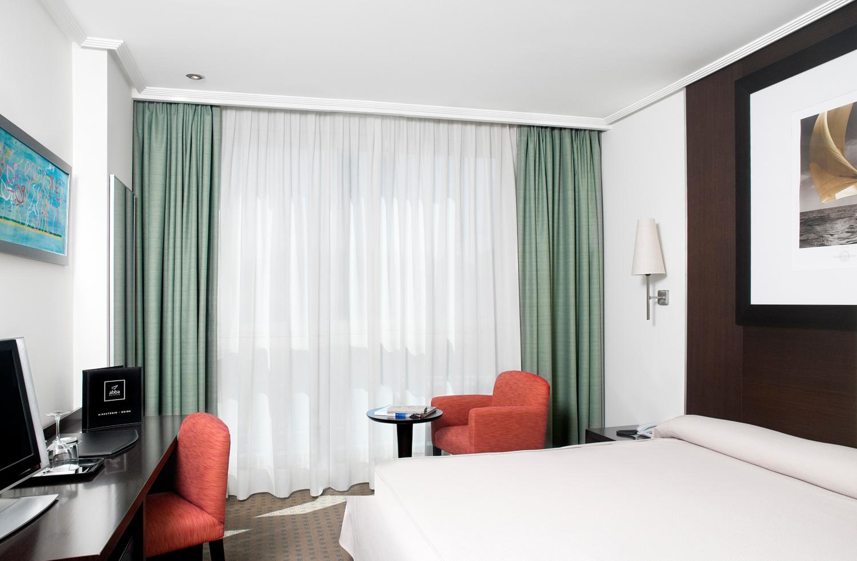 A bedroom featuring a bed with white linens, red armchairs, a dark desk, and pale green drapes.