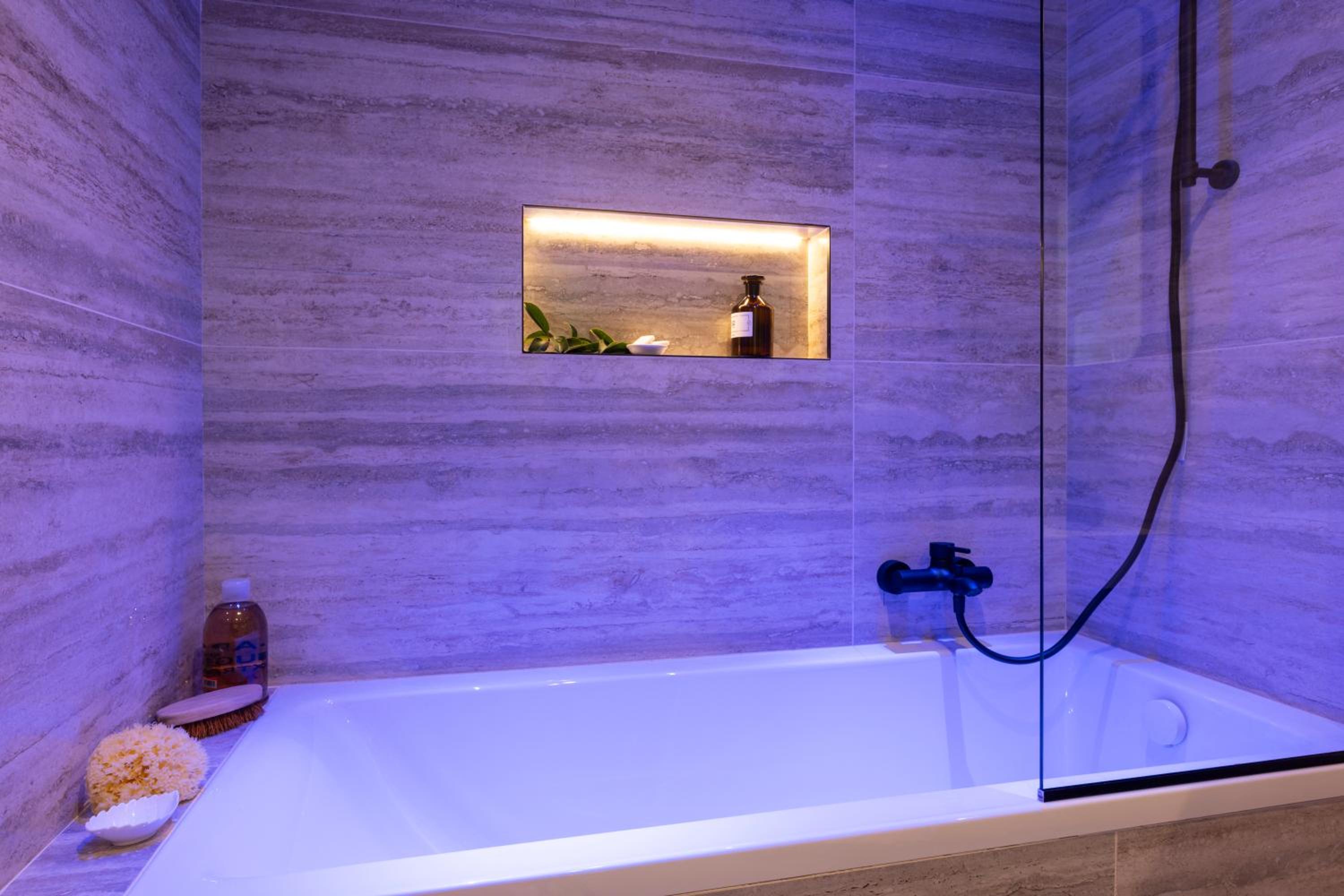 A bathtub beneath textured stone tiles lit in purple, featuring an illuminated wall niche.