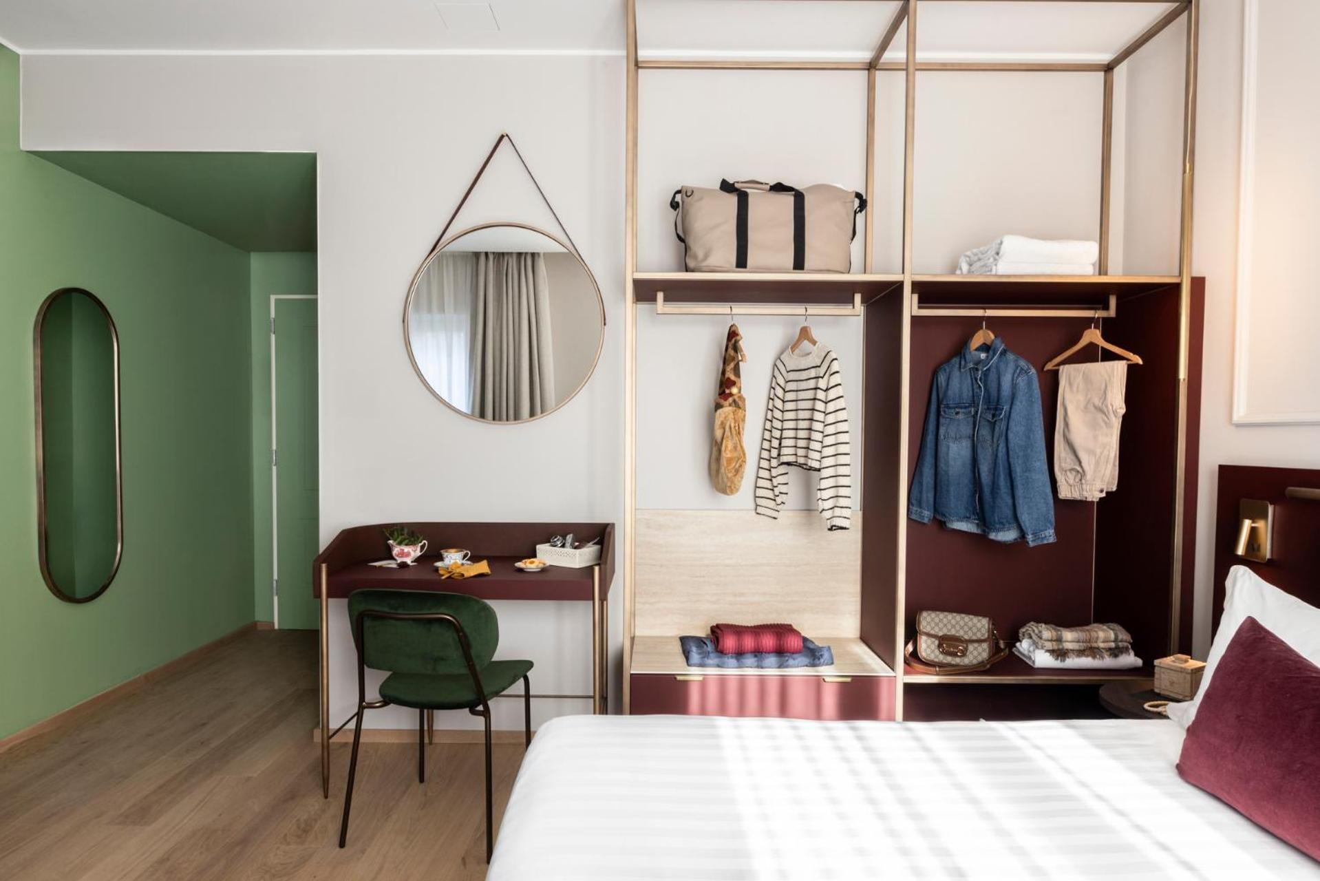 A room corner showing an open wardrobe with hung clothes, a round mirror, small desk, and chair.